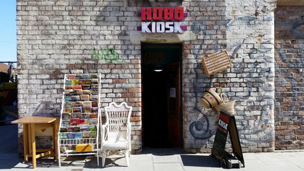 The front entrence of the Hobo Kiosk. a book shelf a collection of books sit in front with a table and chair